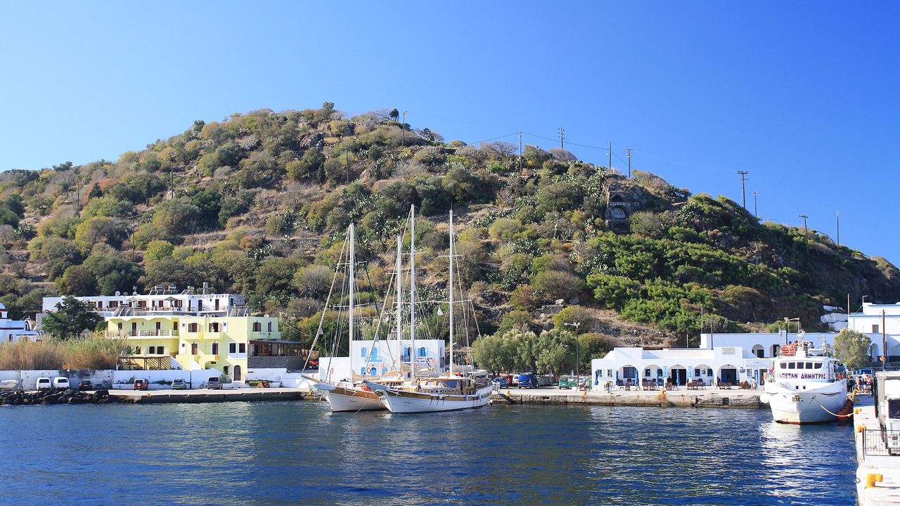 Der Hafen von Mandraki auf Nisyros an einem sonnigen Tag, weiße Häuser und Boote unter einem grünen Hang