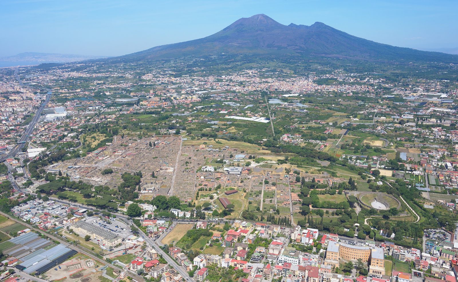 Luftaufnahme der ausgegrabenen Straßen und Häuser von Pompeji bei Tageslicht, dahinter der Vesuv