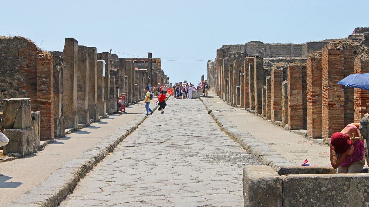 Via dell'Abbondanza in Pompeji: eine lange Straße mit großen antiken Pflastersteinen zwischen erhöhten Gehwegen und zerfallenen Backsteinmauern, in der Ferne Besucher