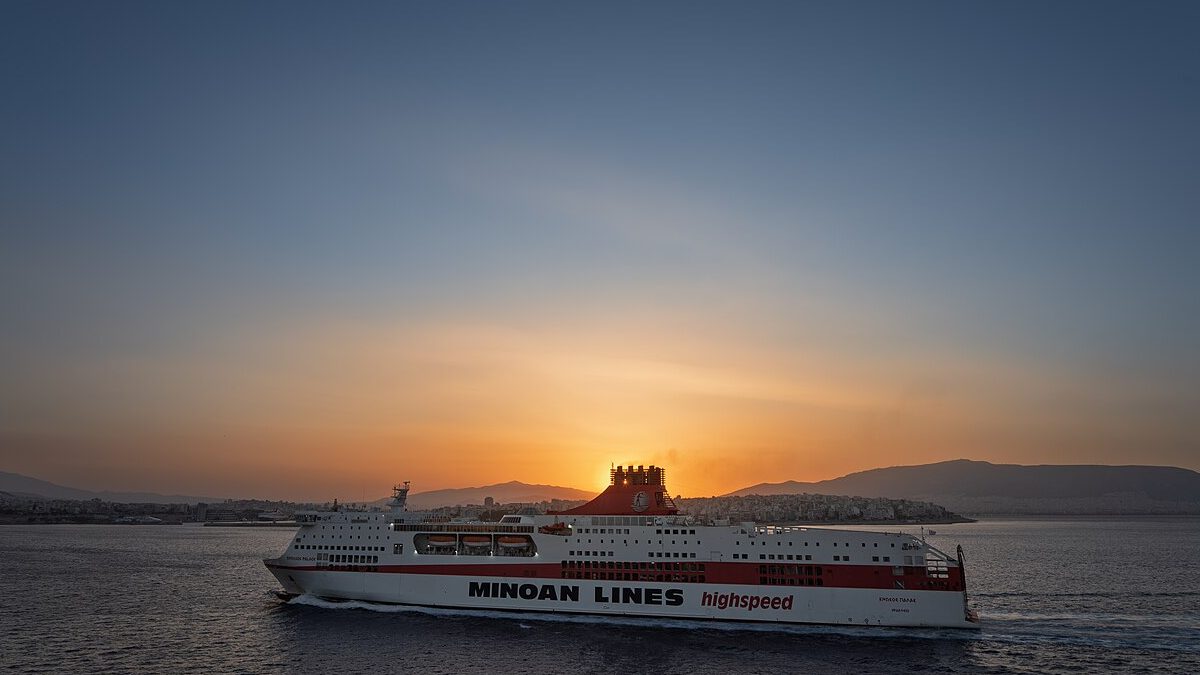 A white and red high-speed ferry leaving Piraeus harbour at sunset, hills along the coast behind it