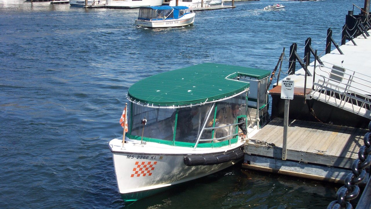 Ein Wassertaxi mit grünem Dach liegt an einem Anleger im Hafen von Boston unter einem Schild für das Flughafen-Wassertaxi, dahinter kreuzt ein weiteres Wassertaxi