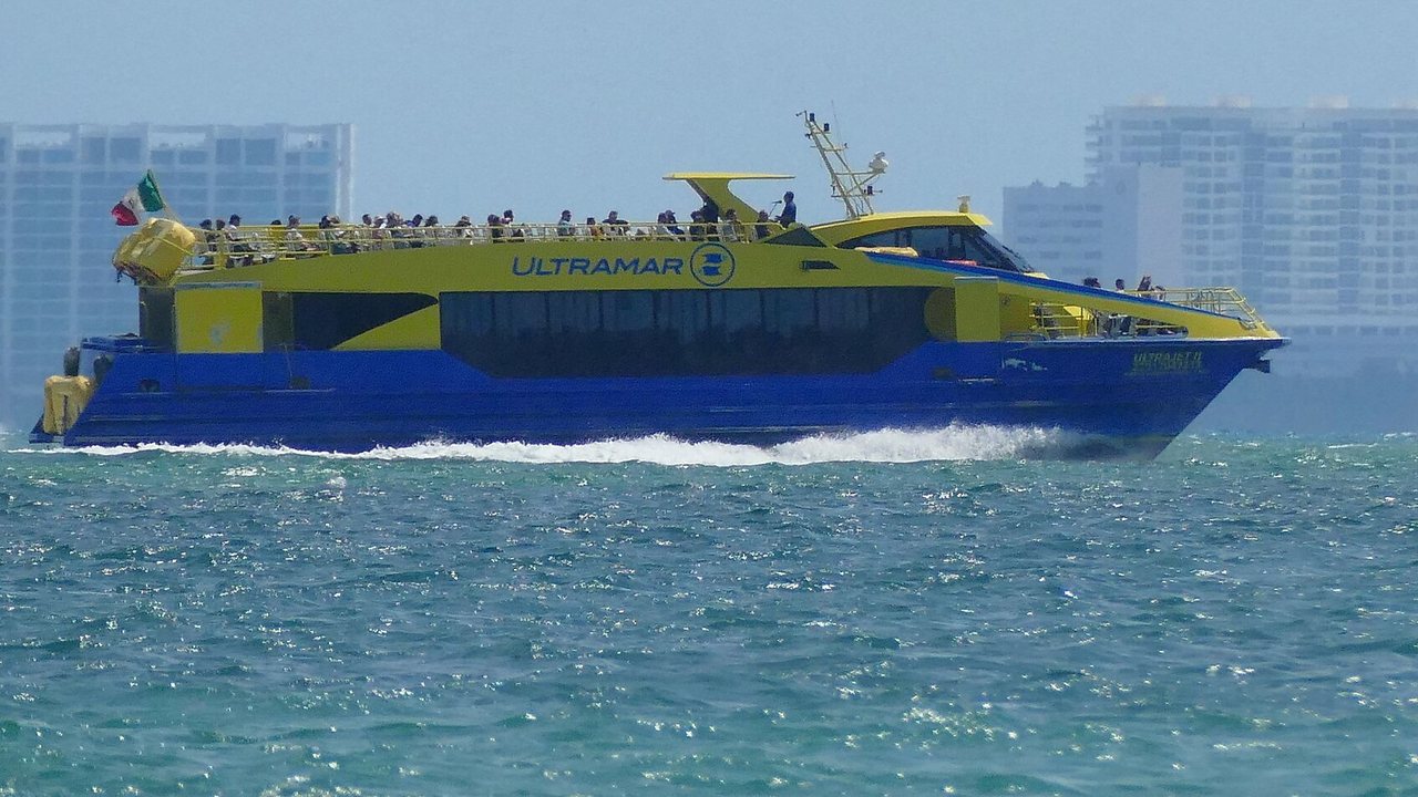A yellow and blue Ultramar fast ferry crossing turquoise water with Cancun hotel towers behind it