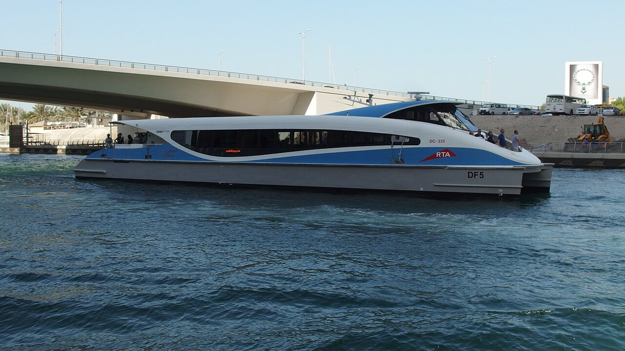 A white and blue RTA Dubai Ferry catamaran on the water in front of a road bridge in Dubai Marina