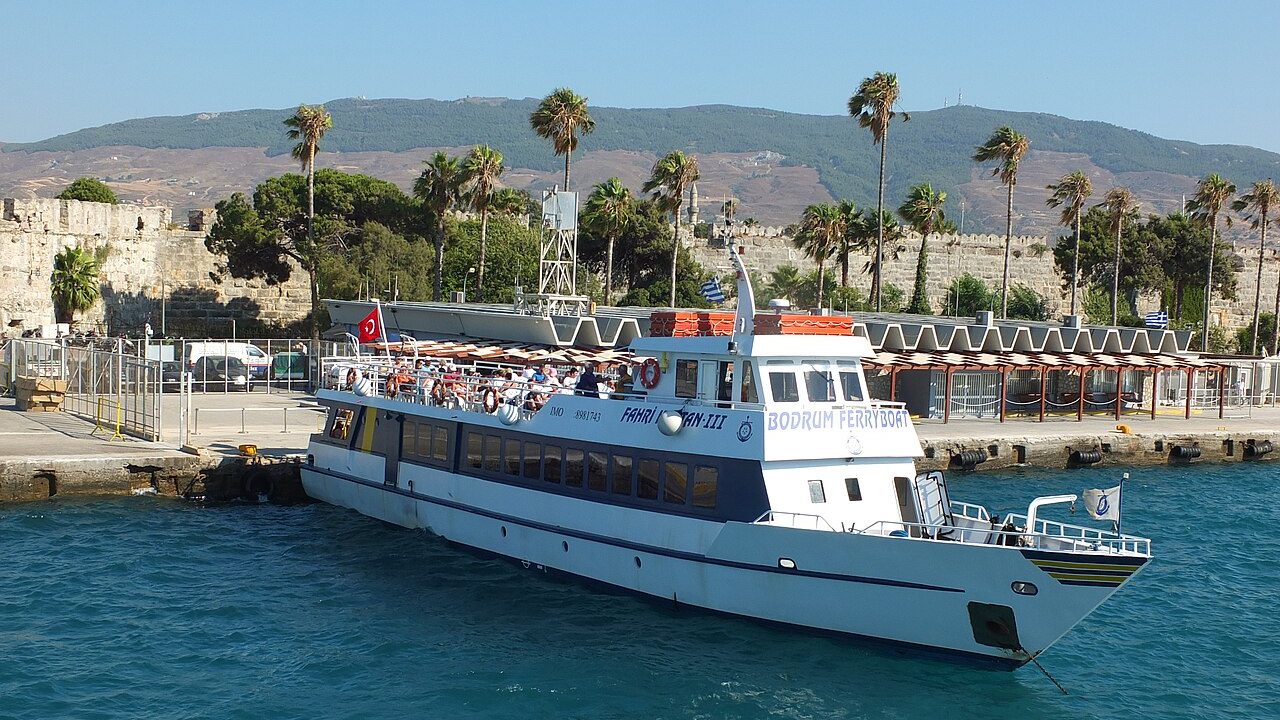 A white passenger boat moored at the quay in Kos harbour, palm trees and the castle walls behind it