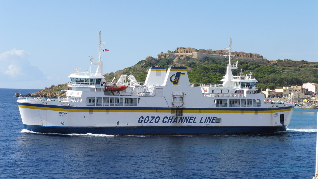 Eine weiße Fähre von Gozo Channel Line mit gelbem Streifen verlässt den Hafen von Mġarr unter einem befestigten Hügel
