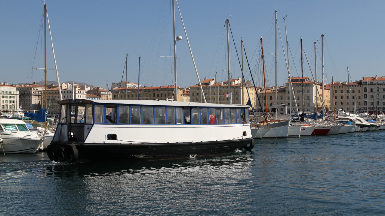 Ein kleines weiß-schwarzes Passagierboot quert den Vieux-Port in Marseille vor festgemachten Yachten und alten Gebäuden