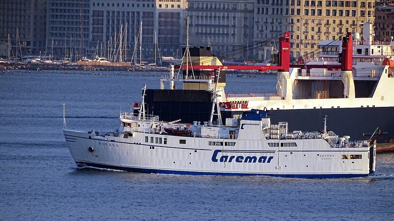 A white Caremar ferry leaving the port of Naples, with the city's waterfront buildings and a larger ship behind it