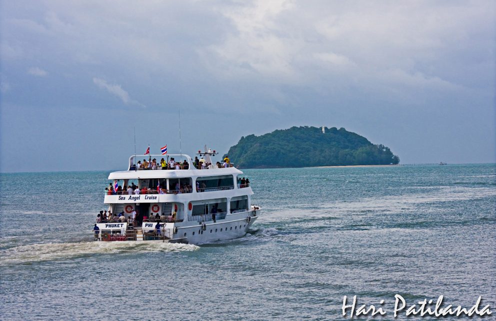 A white passenger ferry with people on its decks heading out to sea from Phuket, a small wooded island behind it under a cloudy sky