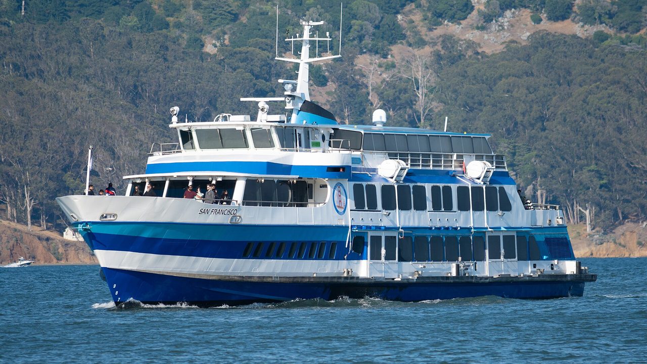 A blue and white Golden Gate Ferry boat named San Francisco crossing the bay in front of wooded hills