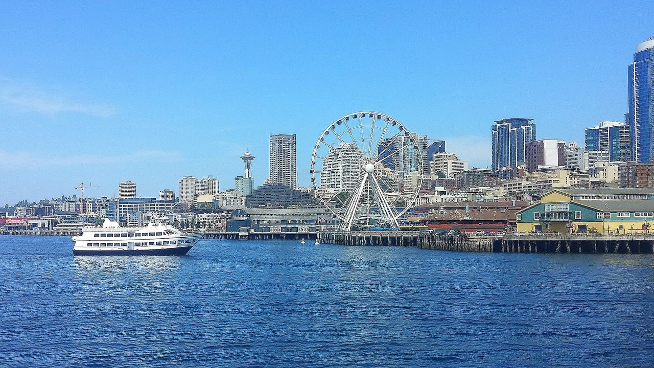 Eine Fähre auf der Elliott Bay vor der Uferpromenade von Seattle mit ihrem Riesenrad an einem sonnigen Tag