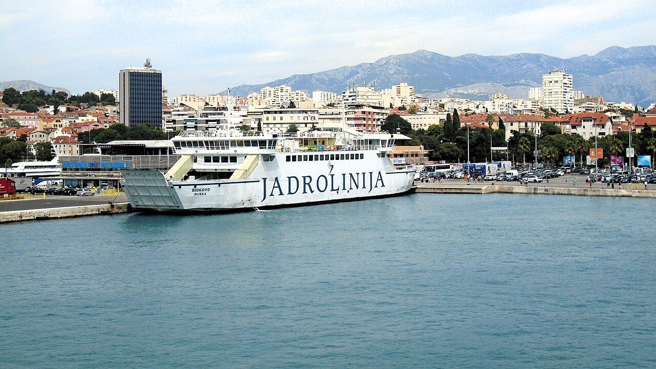 A white Jadrolinija ferry moored at the ferry terminal in Split, the city and hills behind it under a pale sky