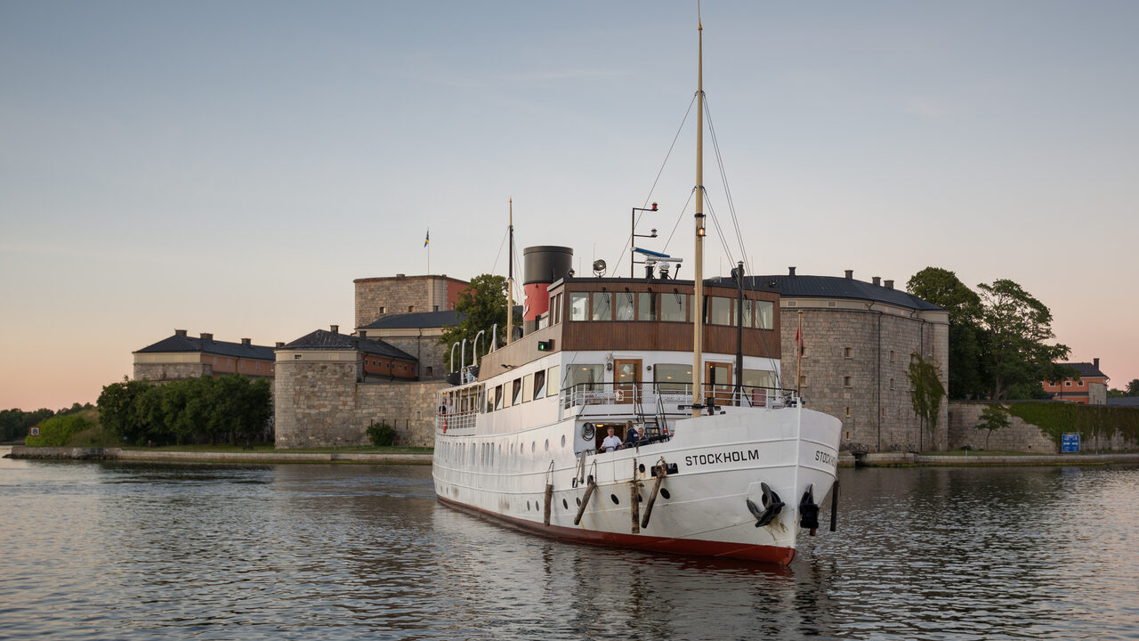 Das weiße Dampfschiff Stockholm auf dem Wasser vor einer grauen Steinfestung in der Abenddämmerung